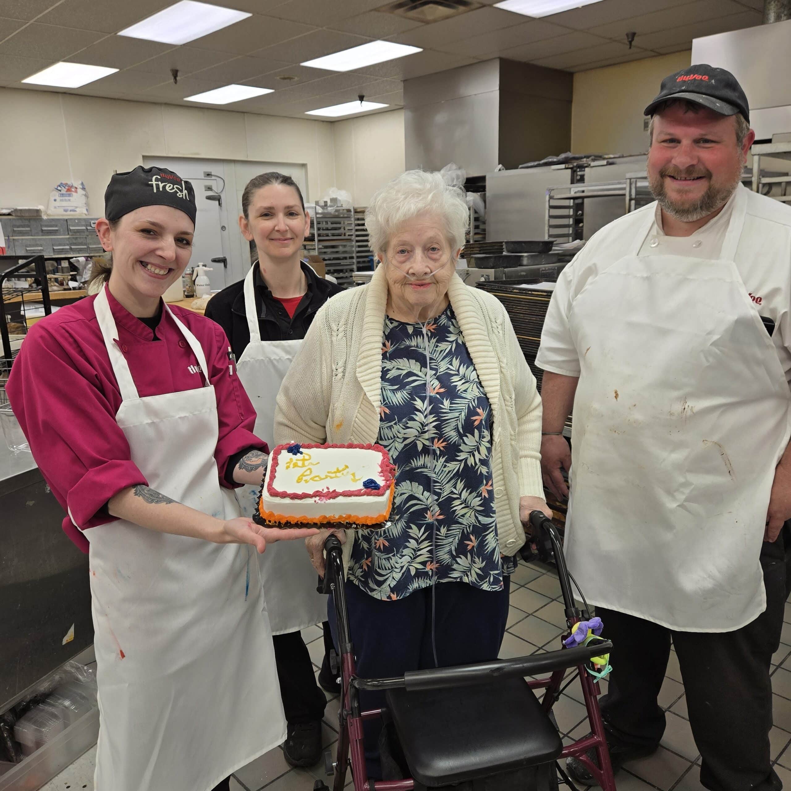 A smiling group of three bakery employees stand with an elderly woman using a walker in a commercial kitchen. The employees, wearing aprons and uniforms, present a small decorated cake that the elderly woman holds at the center of the group.