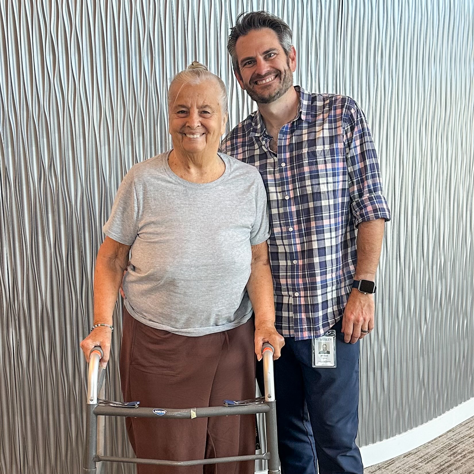 Women smiling while holding a walker. Man standing beside her, smiling, with one arm wrapped around her.