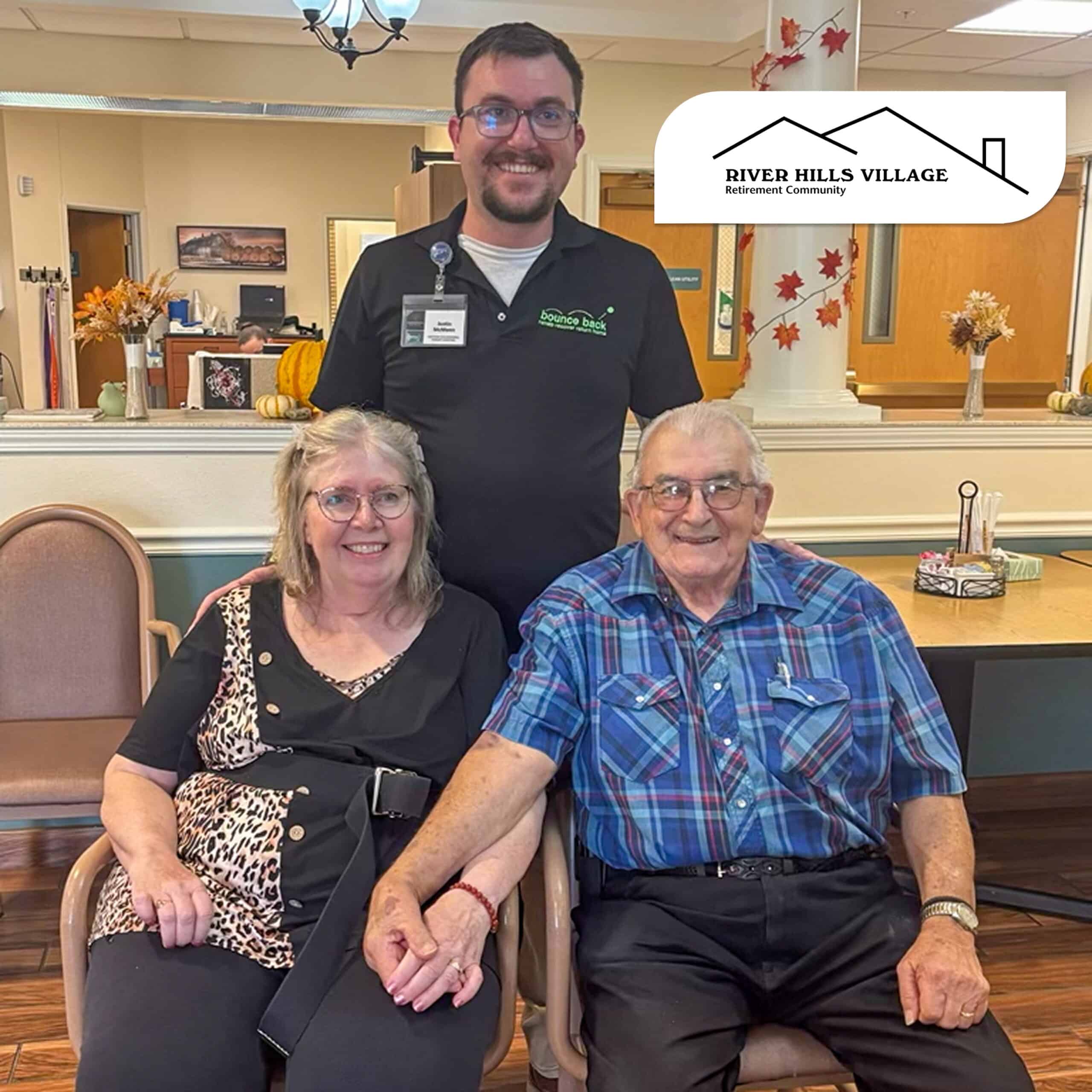 Three people smiling at a retirement community. An elderly couple sits holding hands in chairs, with a staff member standing behind them. Autumn decorations are visible in the room, and a ‘River Hills Village Retirement Community’ logo appears in the upper right corner.