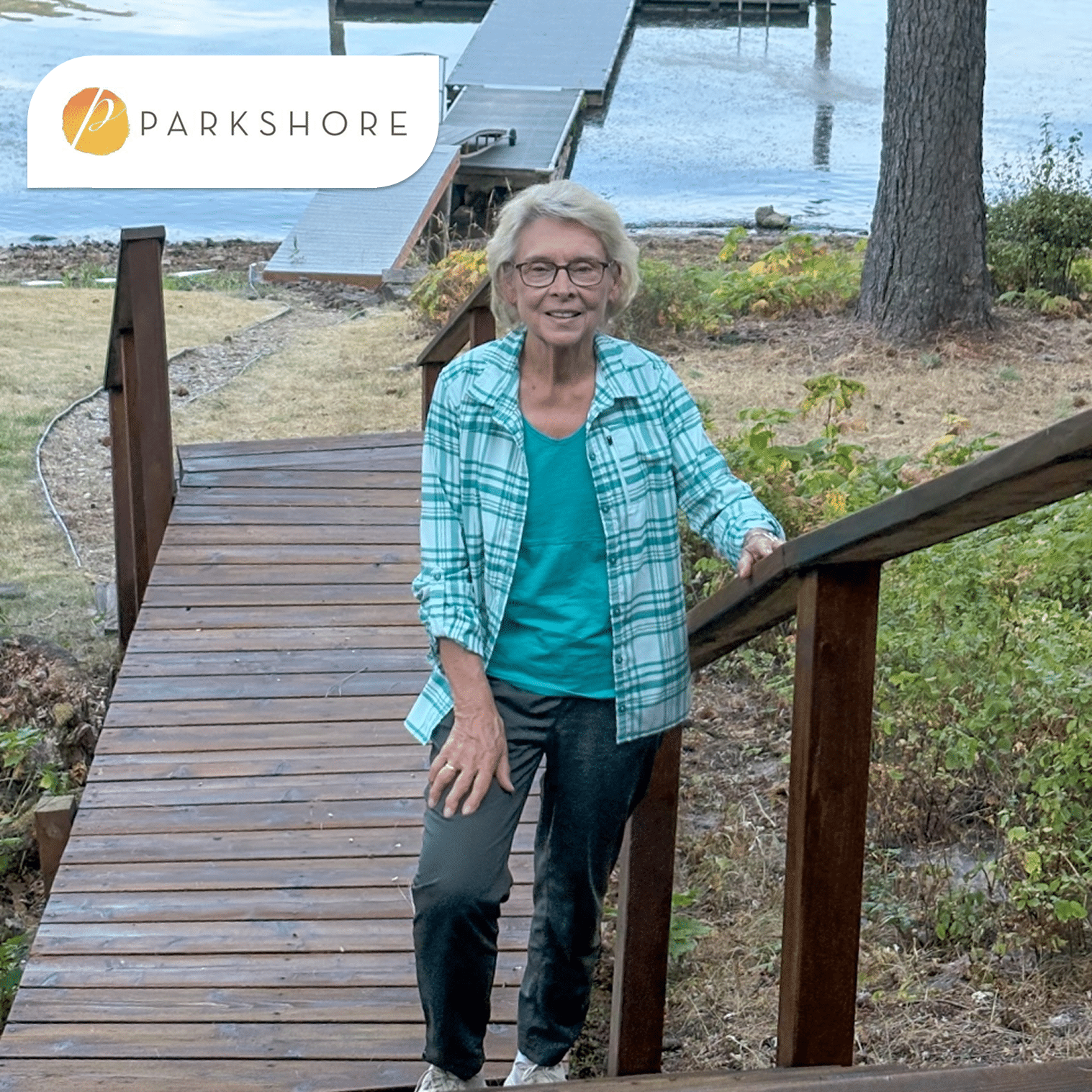 An older woman with short gray hair and glasses stands on a wooden ramp by a lakeside dock, wearing a teal shirt and green plaid overshirt, with trees and water in the background.