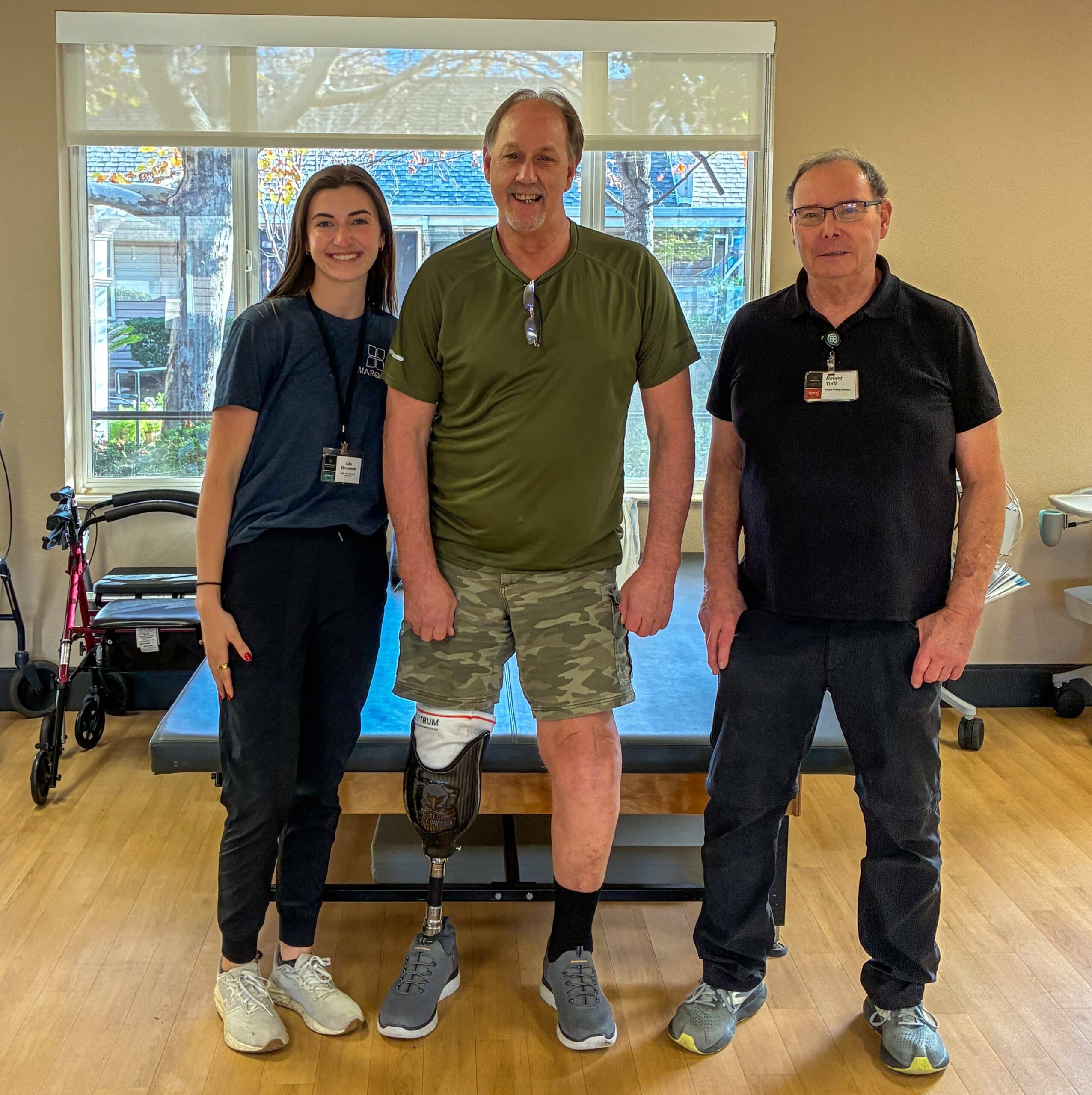 Three people stand side by side in a physical therapy or rehab room. The person in the center is a smiling man wearing a green shirt, camo shorts, and a left-leg prosthetic, standing confidently. He is flanked by two staff members wearing name badges and athletic clothing. A therapy table and a walker are visible behind them, with large windows letting in natural light.