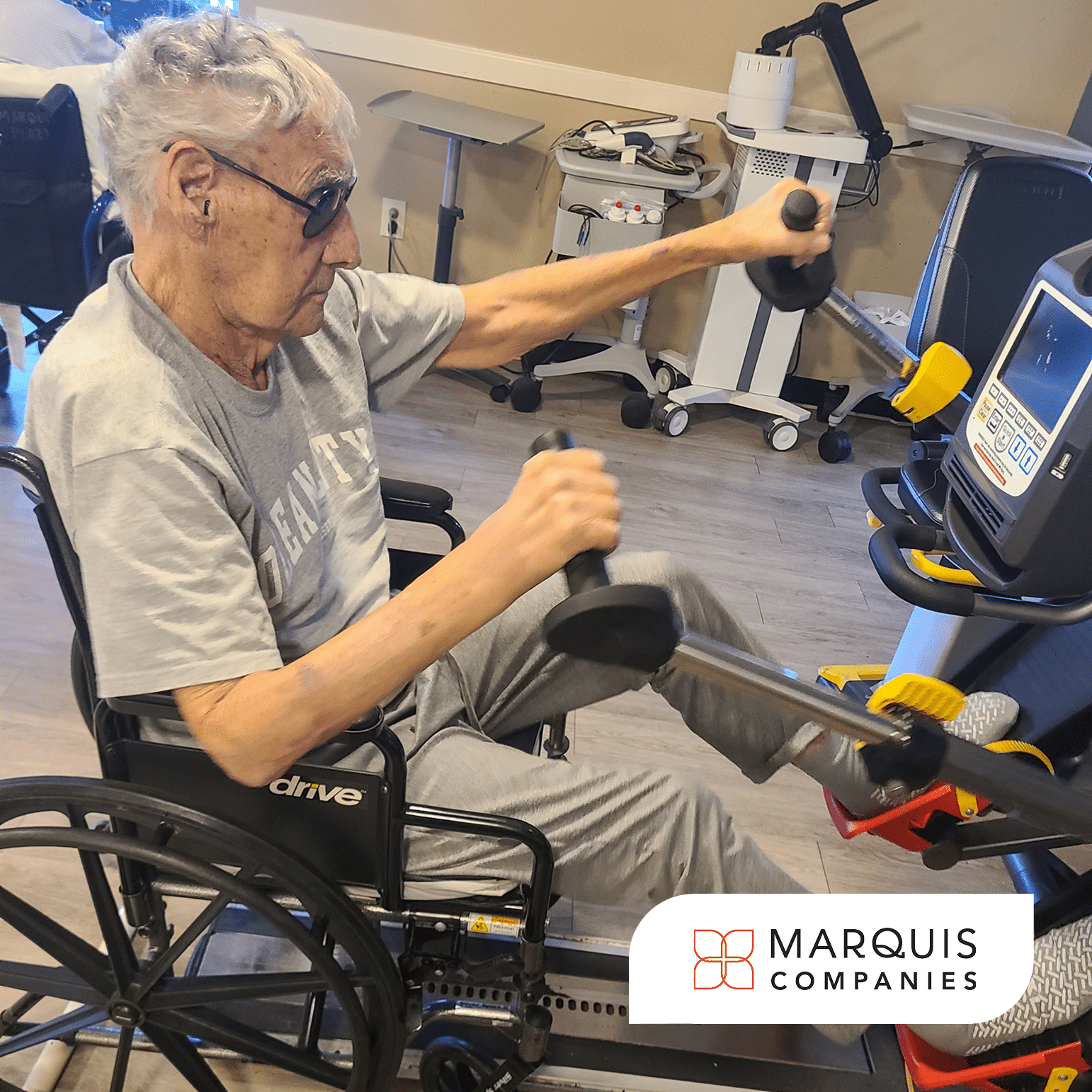 A resident exercises in a therapy gym while seated in a wheelchair, using a seated exercise machine and holding hand weights. Focused on his workout, he works to rebuild his strength and endurance as part of his rehabilitation.