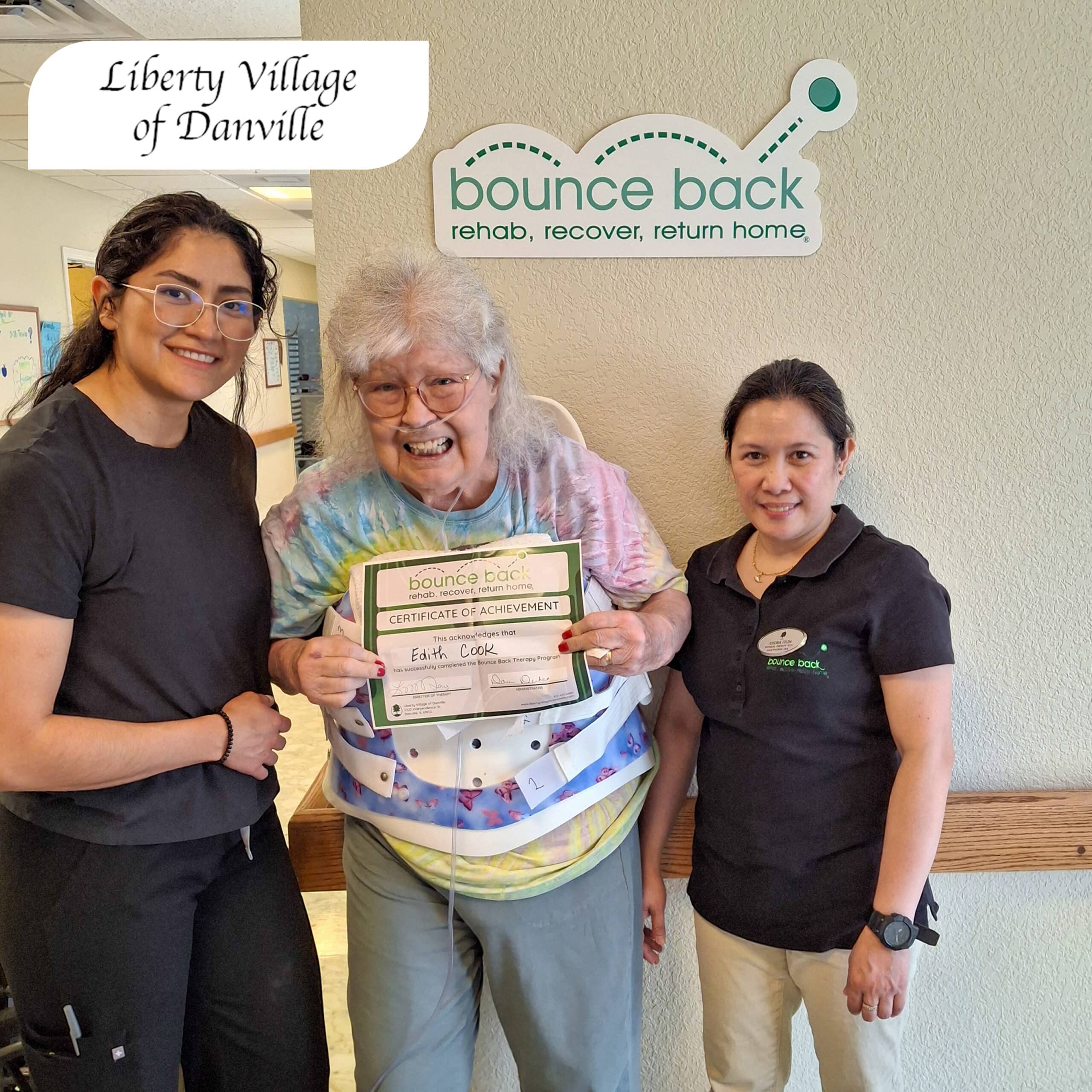 An older woman wearing a tie-dye shirt and a supportive back brace smiles while holding a Bounce Back Certificate of Achievement. She stands between two rehabilitation team members beneath a Bounce Back Therapy sign at Liberty Village of Danville, celebrating her recovery progress.