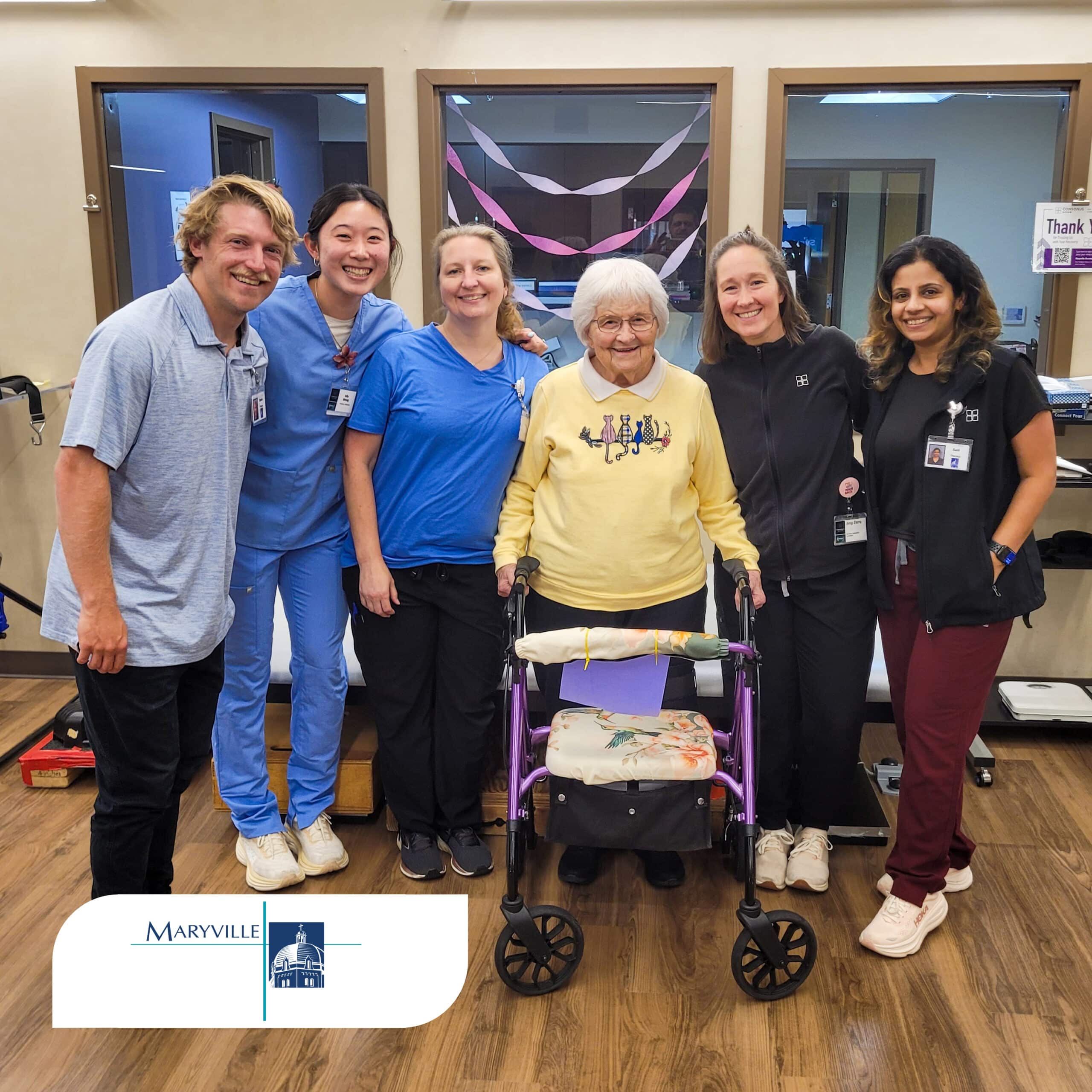 An older woman wearing a yellow sweater stands smiling behind a purple walker while surrounded by five smiling rehabilitation team members in a therapy gym. Pink and white streamers decorate the windows behind the group, and a Maryville logo appears in the bottom left corner of the image.