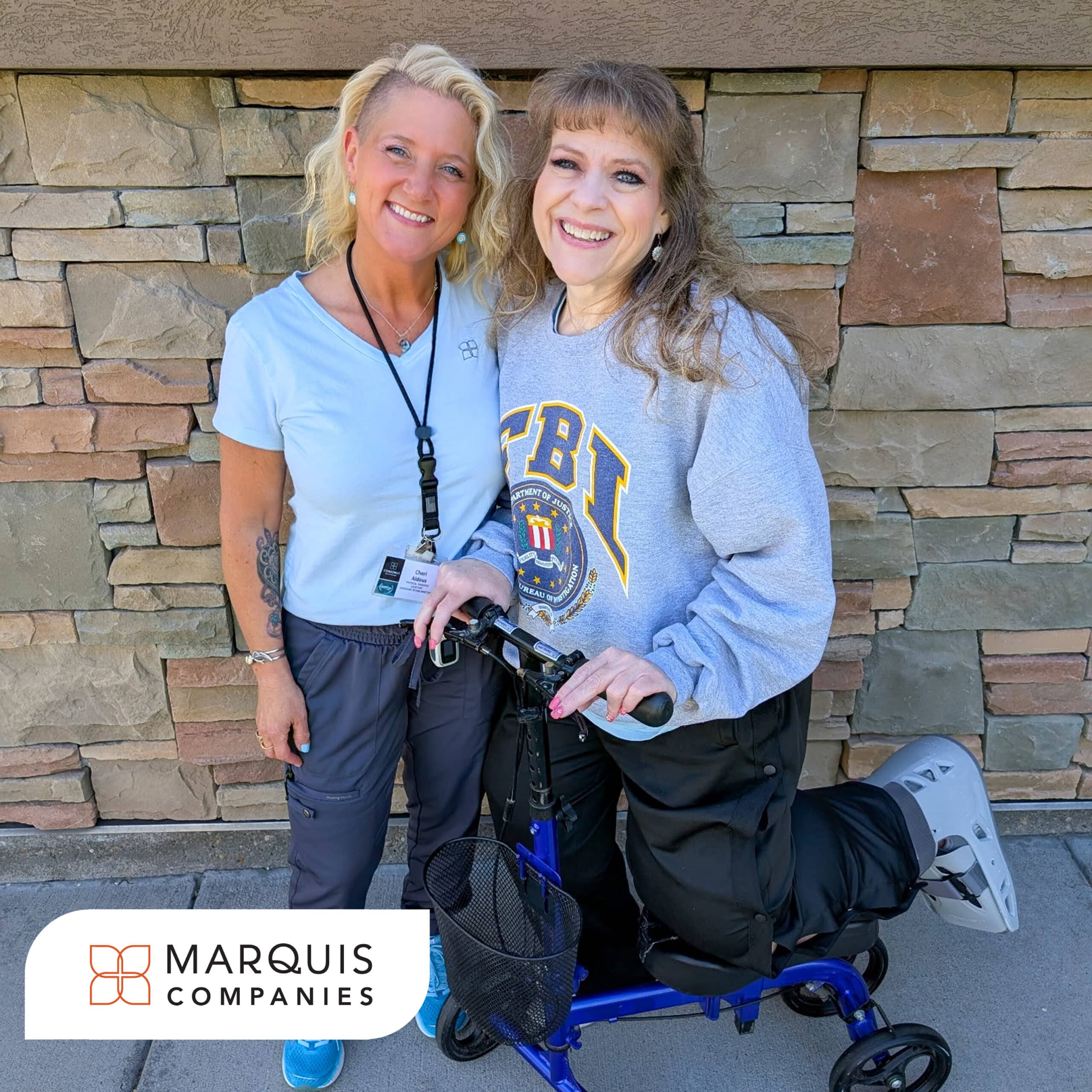 A smiling woman using a blue knee scooter poses beside a rehabilitation team member outside in front of a stone wall. The woman wears an FBI sweatshirt and a walking boot on one leg, while the staff member stands with her arm around her. A Marquis Companies logo appears in the bottom left corner of the image.